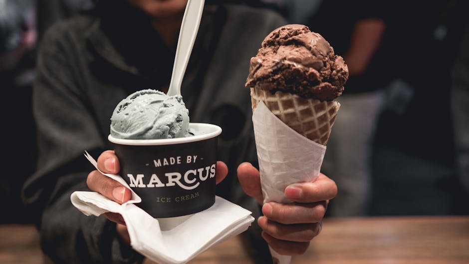 The image features a person holding two types of ice cream: in the left hand, a small black paper cup labeled 'Made by Marcus Ice Cream' filled with light grayish-green ice cream, accompanied by a whi